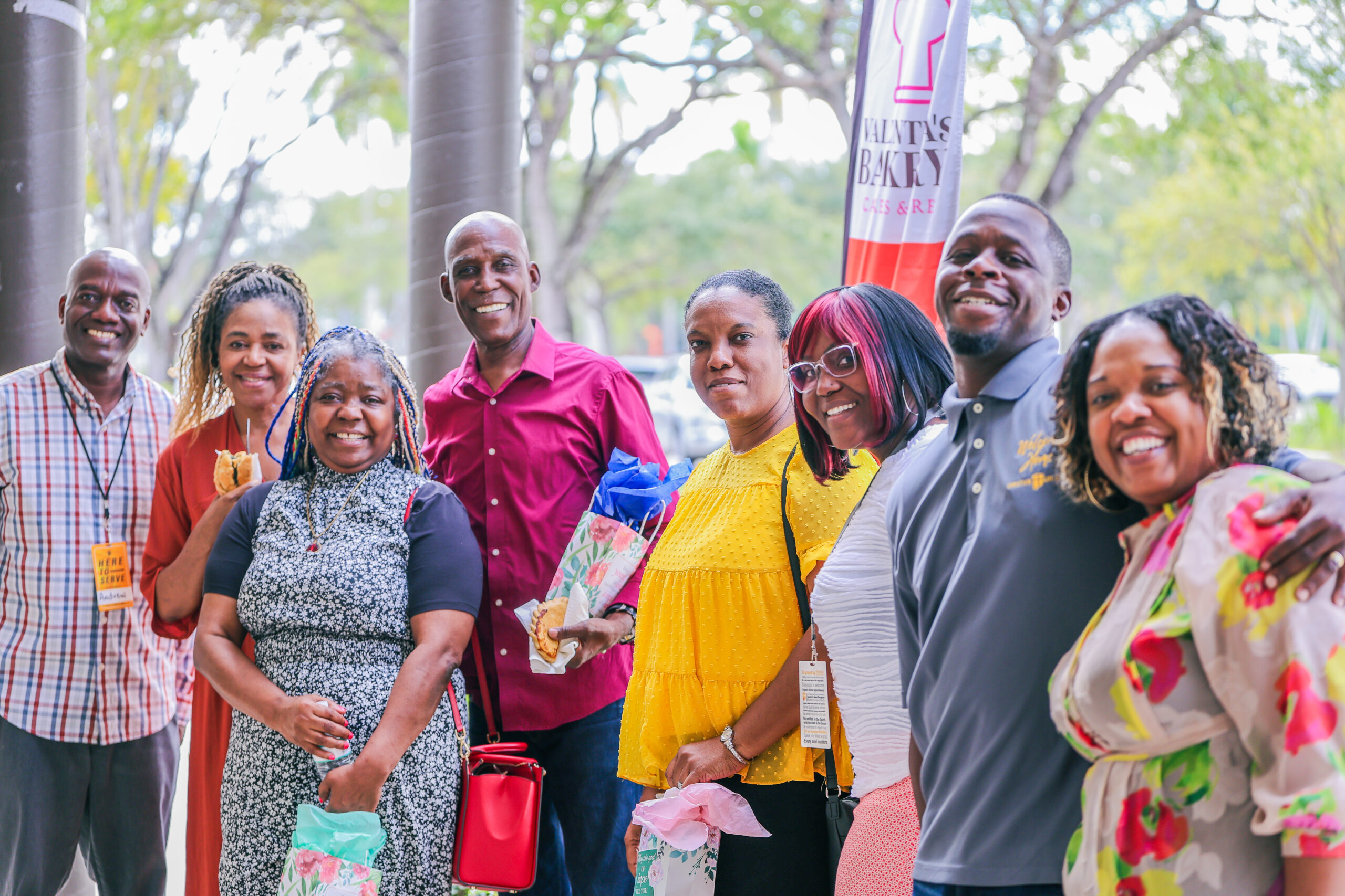 The photo shows a group of people smiling and posing together outdoors, holding gift bags and food, with a bakery banner visible in the background.