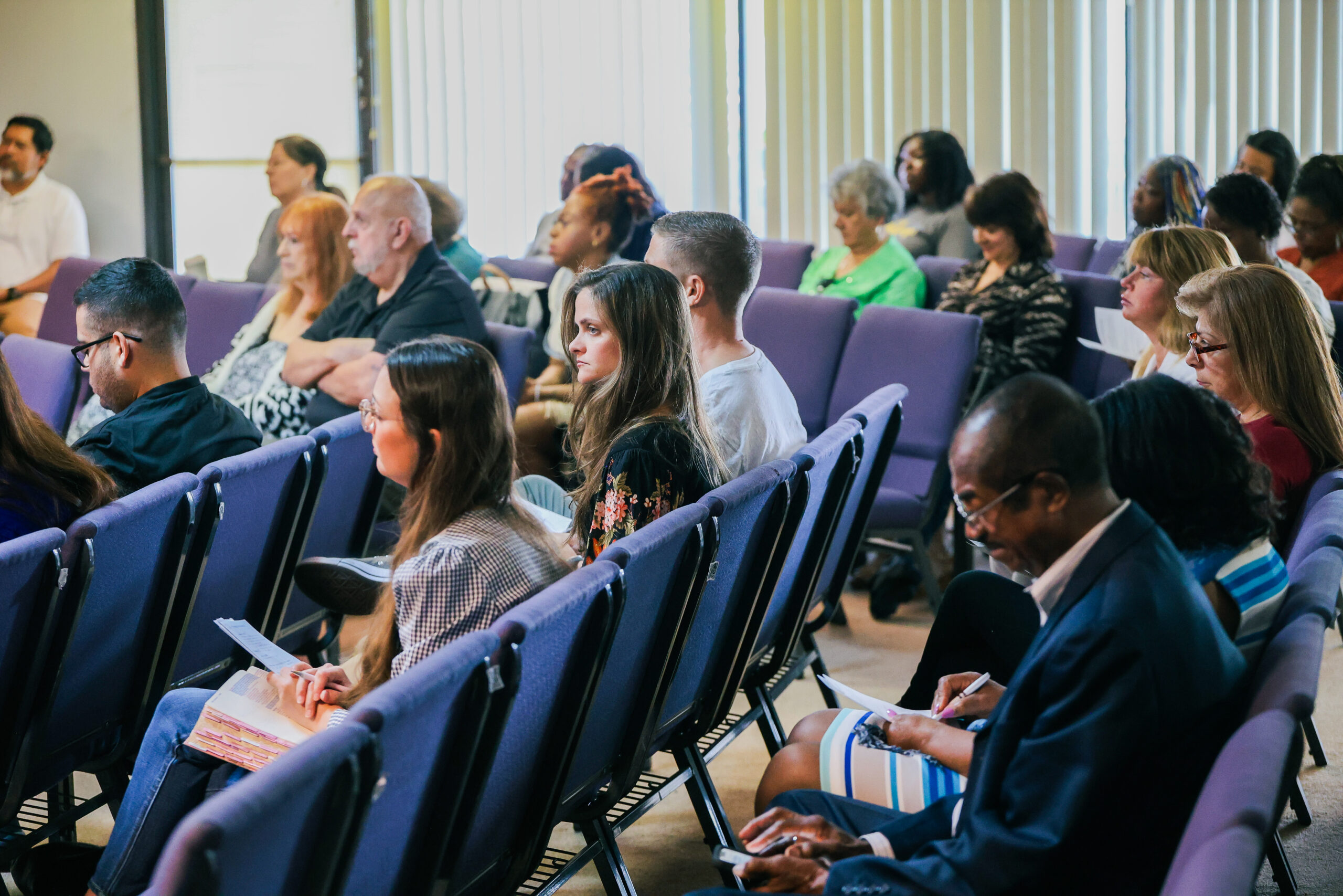 The photo shows a diverse group of people seated in rows of purple chairs, attentively listening in an indoor setting with natural light streaming through vertical blinds.