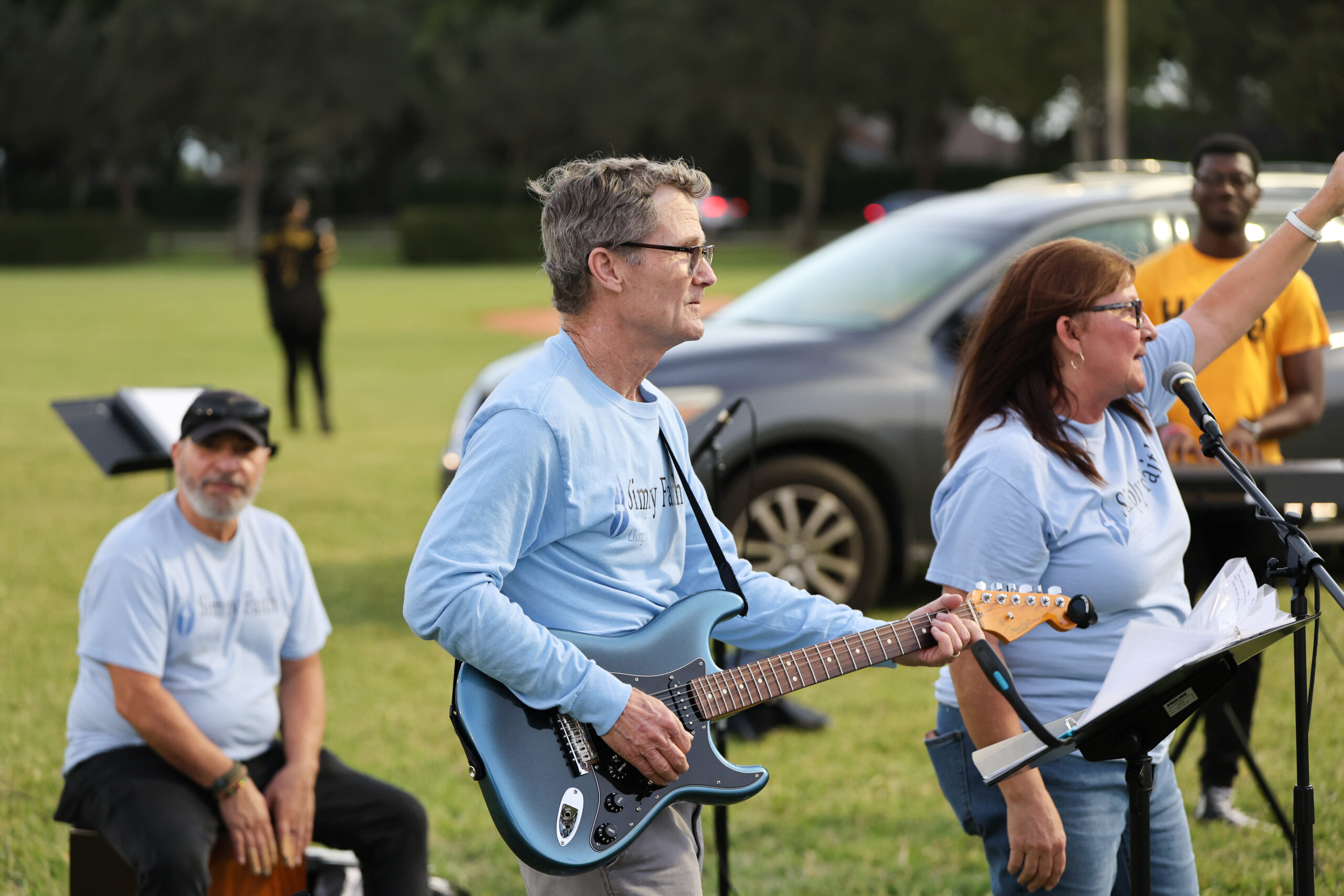 The image shows a group of musicians and speakers in a park, with a man playing guitar and a woman addressing the crowd, both in matching blue shirts.